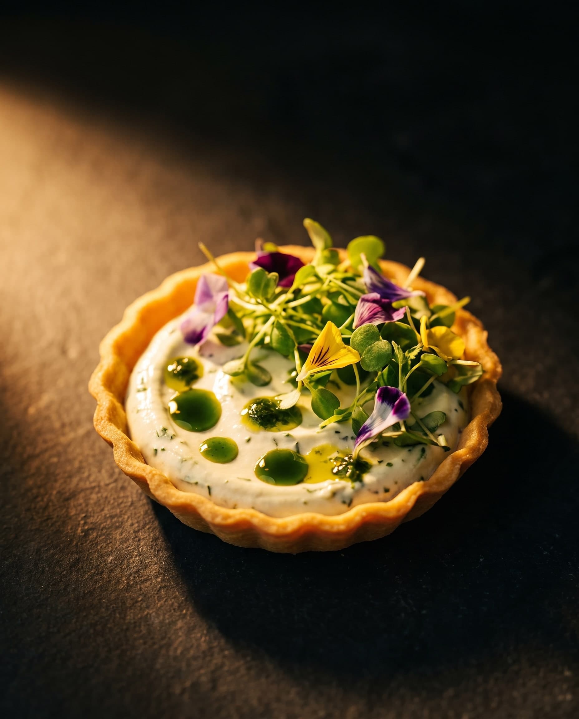 Tartelette mit heller Kräutercreme, Kresse, essbaren Blüten in Violett und Gelb und Tupfen von Kräuteröl, im warmen Streiflicht auf dunklem Stein